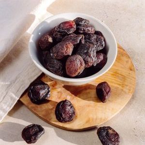 High-quality photo of dried prunes in a white bowl, set on a wooden board with natural lighting.